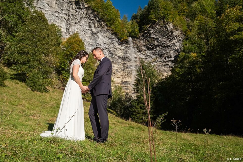 Reportage de mariage en automne à la montagne : portrait des mariés sous la falaise et sa cascade en sépia