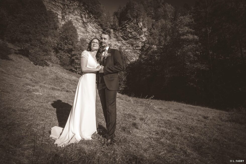 Reportage de mariage en automne à la montagne : photo des mariés sous la falaise et sa cascade en sépia